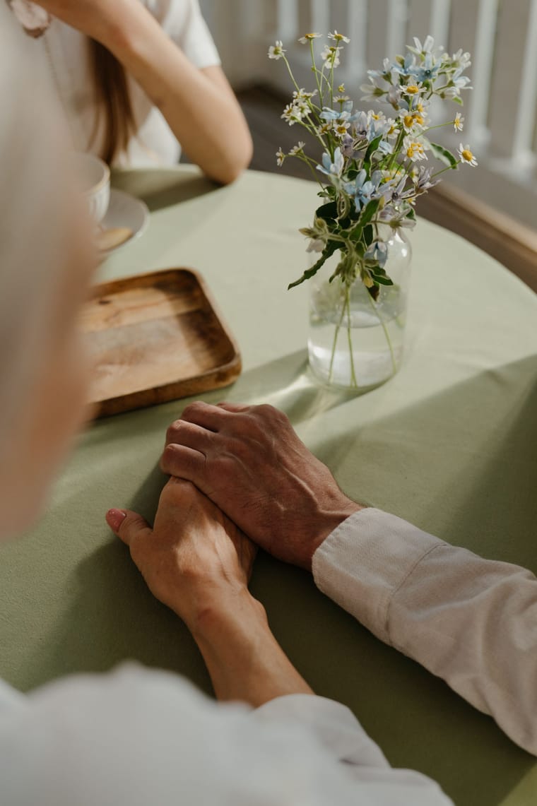 An older hand and a younger hand clasped together on a sage-green table with wildflowers.